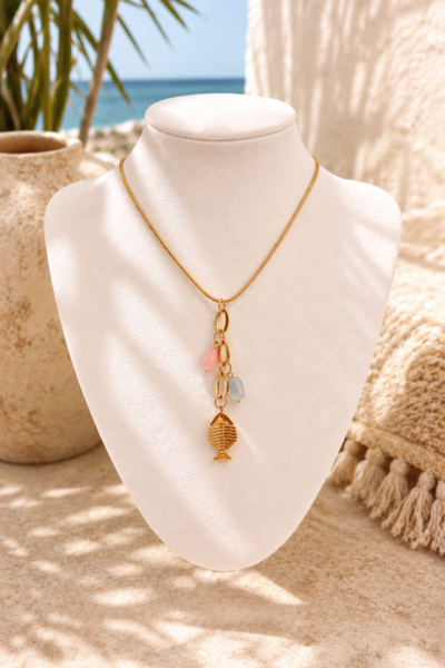 The ocean whisper necklace Gold toned stainless steel necklace with fish charm and pastel pink and blue details displayed in an Ibiza inspired beach setting