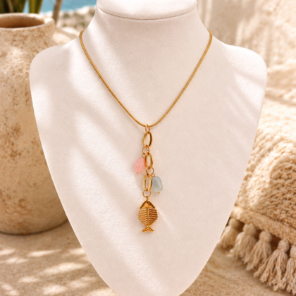 The ocean whisper necklace Gold toned stainless steel necklace with fish charm and pastel pink and blue details displayed in an Ibiza inspired beach setting