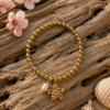 Pearl Bloom Bracelet β Gold Stainless Steel Gold stainless steel beaded bracelet with freshwater pearl charm and flower pendant displayed on driftwood with cherry blossom accents.